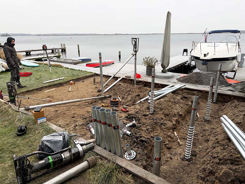 Schroefpalen en materieel op bouwlocatie voor vlonder aan IJsselmeer, met boot en steiger op de achtergrond